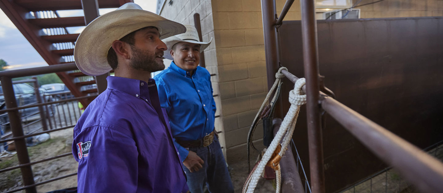 Dustin Boquet wearing a purple shirt and a hat talking with a friend wearing a blue shirt before the rodeo.